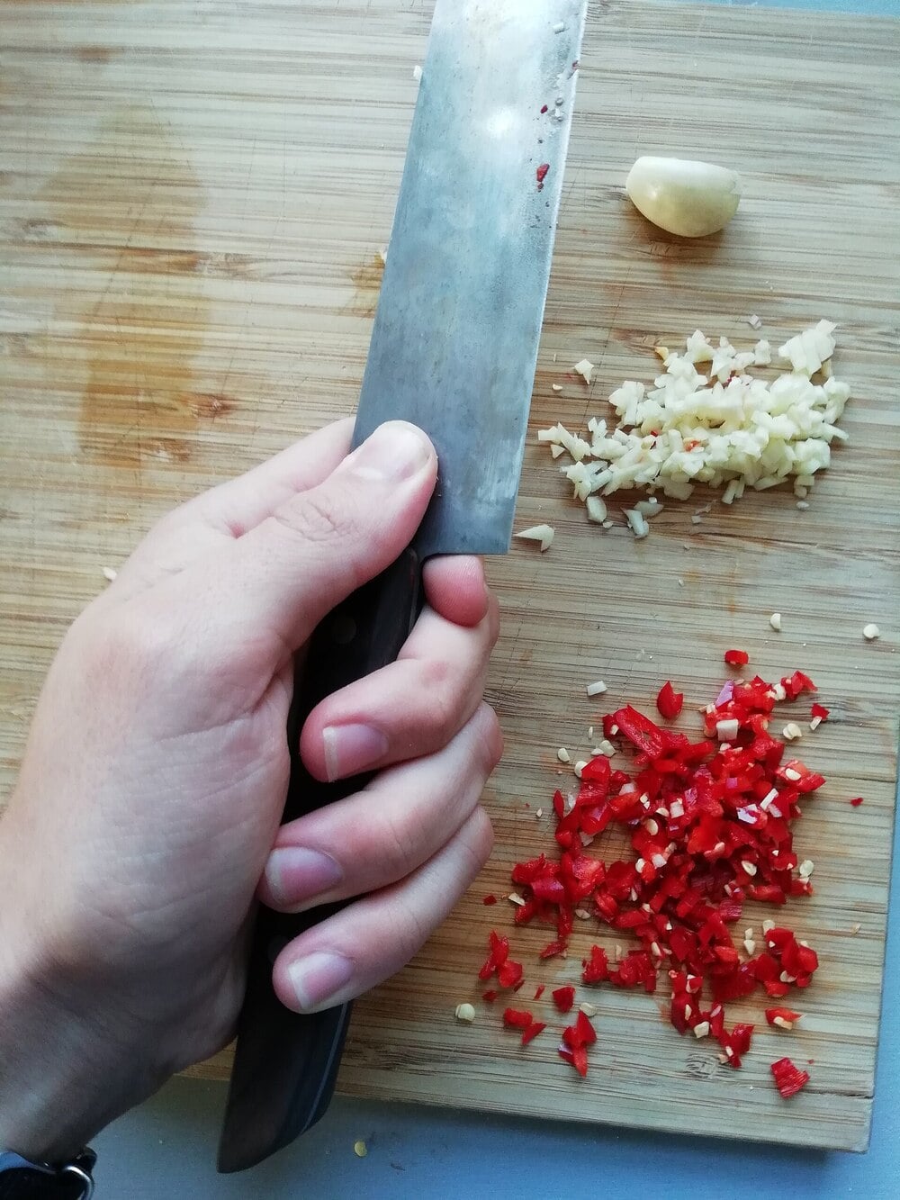 A hand holding a knife above a wooden cutting board with chopped garlic, chopped red chili, and a garlic clove.