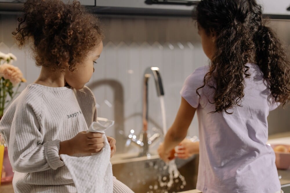 Two young children stand at a kitchen sink; one washes dishes while the other dries with a towel. Water splashes from the tap as they work together.