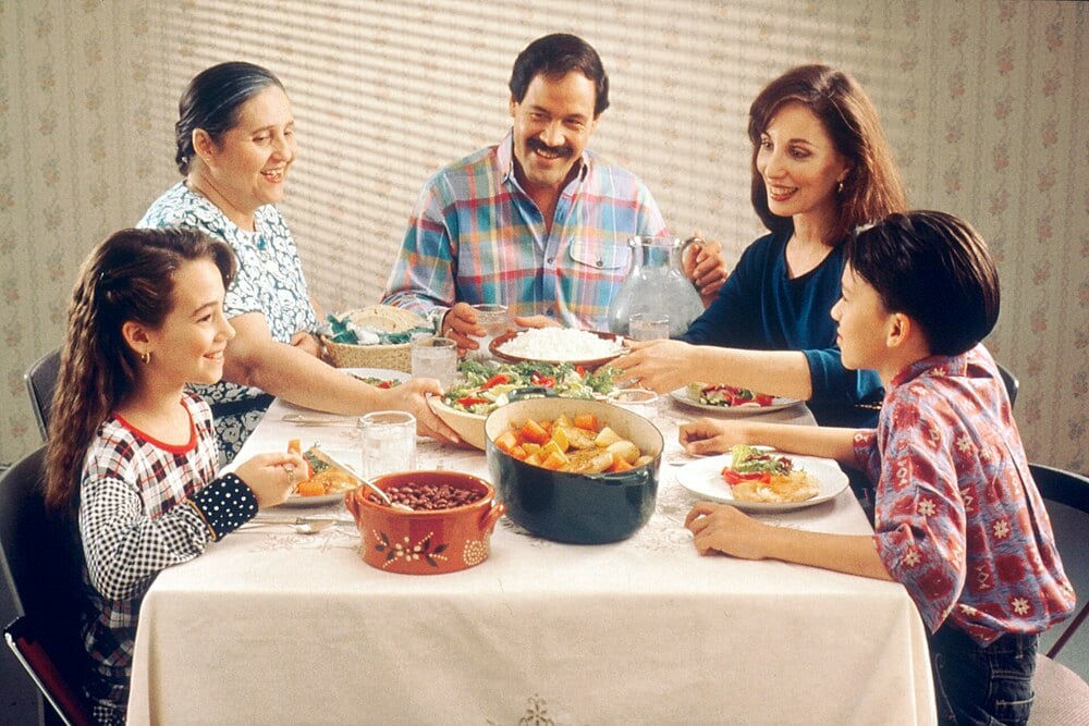 A family of five sits at a table, smiling and sharing a meal. The table is set with dishes like salad, beans, vegetables, and water. Sunlight shines through blinds onto the cheerful scene.