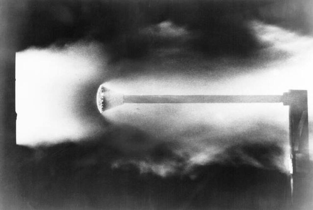 A high-speed photograph shows a bullet mid-flight after being fired, with smoke and shock waves visible around it against a dark background.