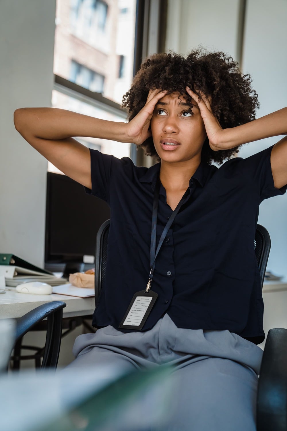 A woman sits at a desk with her hands on her head, looking stressed or overwhelmed. She wears a black shirt and ID badge, with office equipment and windows in the background.