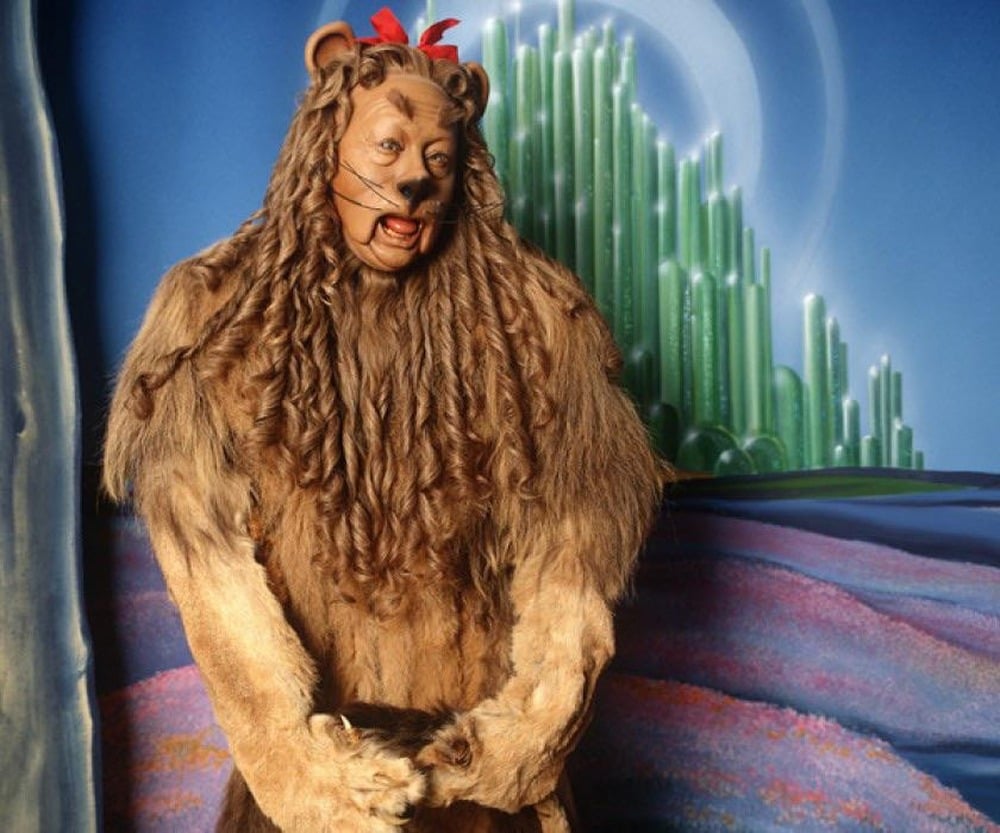 A person in a lion costume with a red bow stands in front of a colorful background featuring a green castle and a blue sky, reminiscent of The Wizard of Oz.