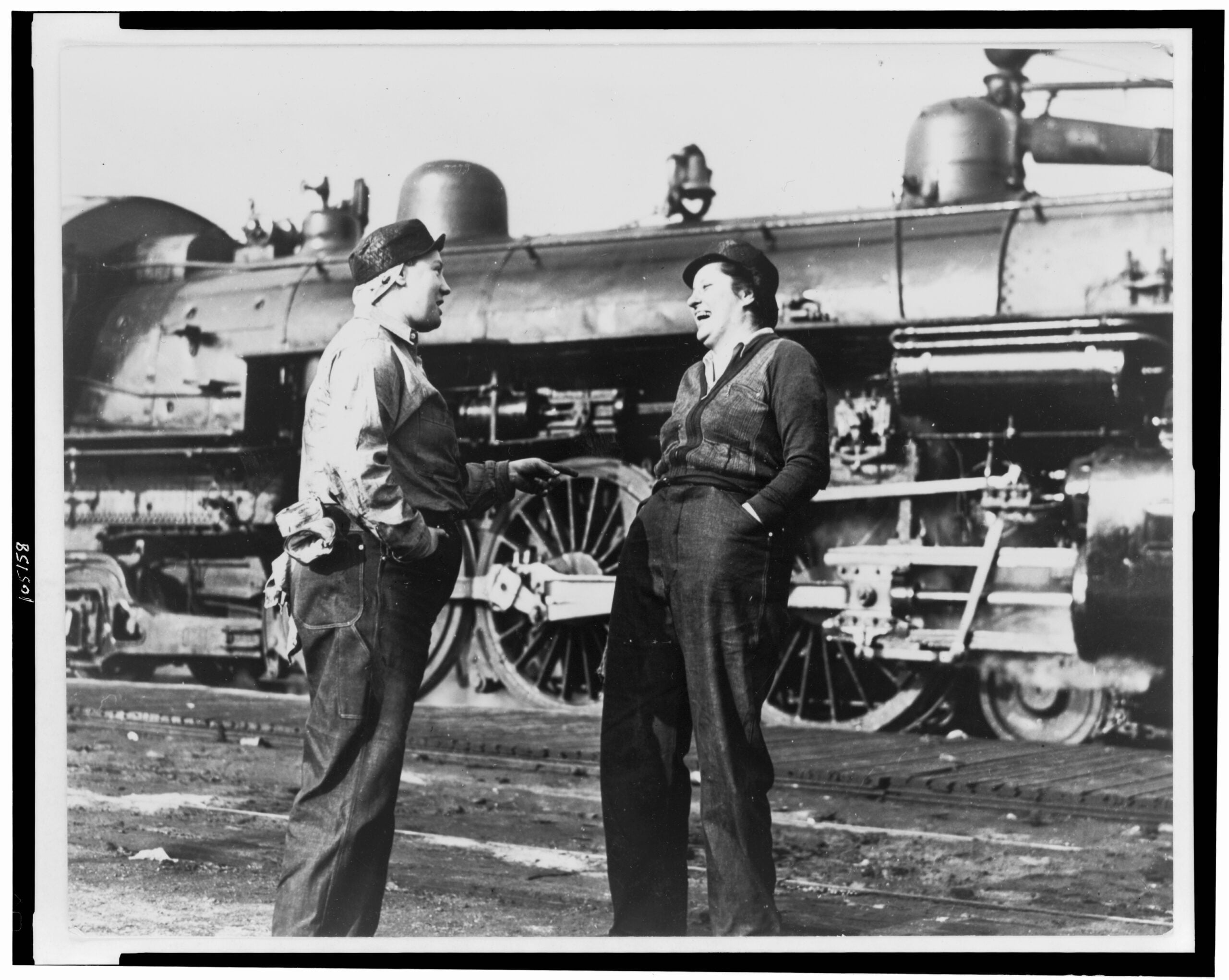 Two people in work clothes stand laughing and talking in front of a large steam locomotive on train tracks, with machinery details visible in the background.