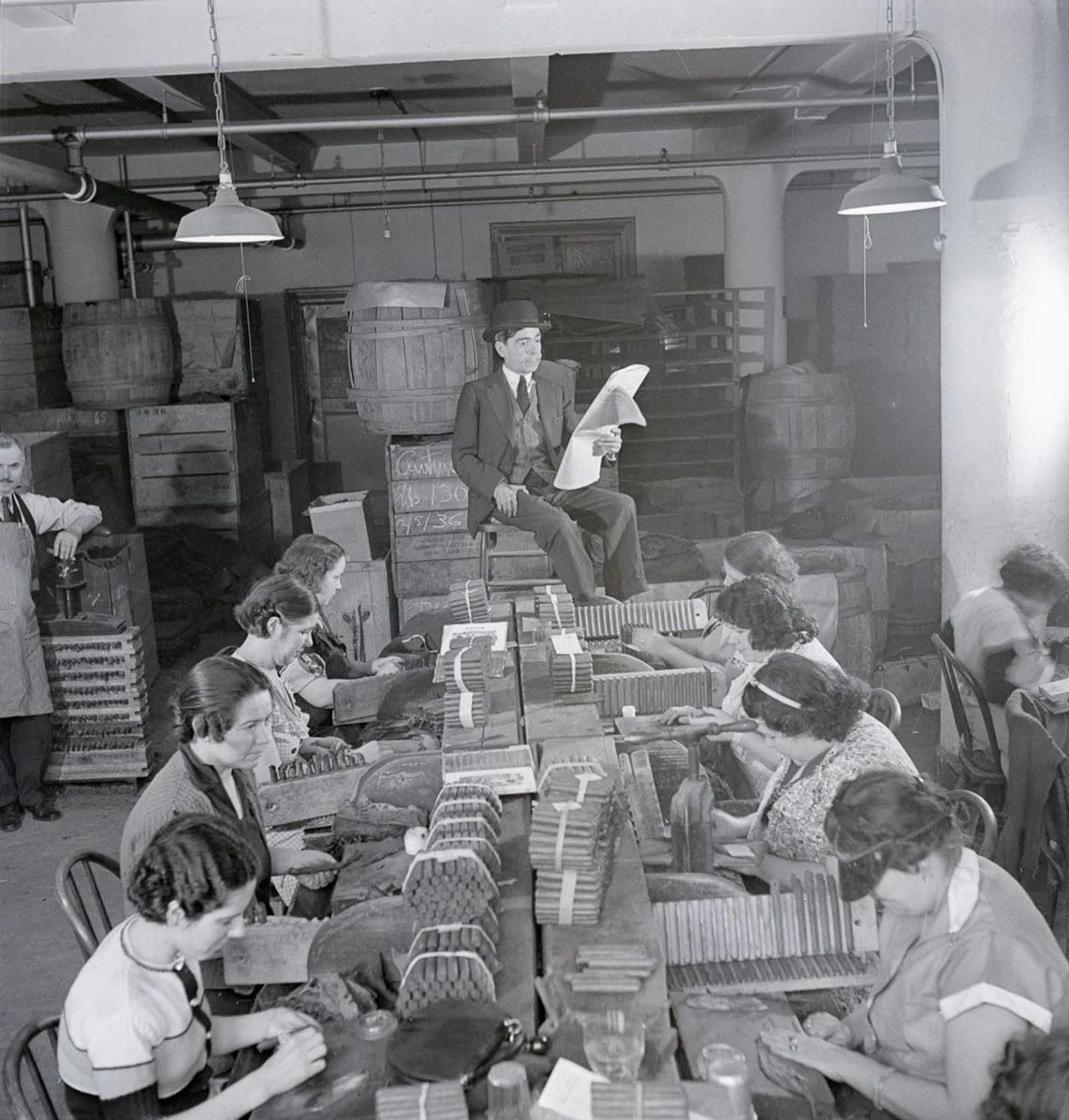 Workers sit in rows making cigars at long tables, while a man in a suit sits elevated at the end, reading from a paper. The room is filled with barrels and lit by overhead lamps.