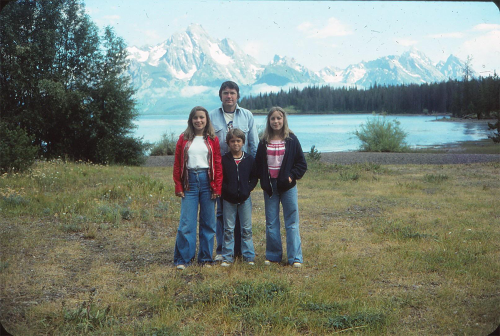 A man and three children stand on grass near a lake, with tall mountains and trees in the background. All are wearing jackets or sweaters and jeans, and they are smiling at the camera.