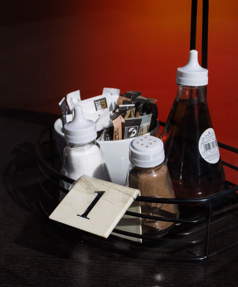 A tabletop caddy holds sugar packets, two shakers with white and brown powders, and a bottle of syrup. A table number card with the number 1 is placed in front. The background is dark with a gradient of red.
