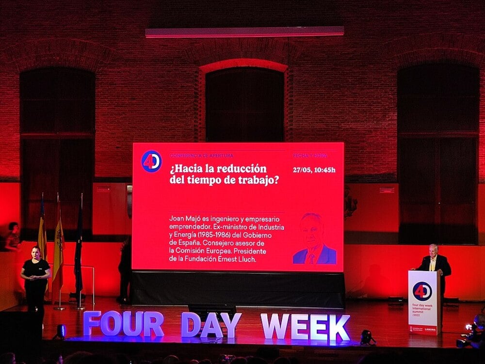 A speaker presents at a conference with a large red screen behind him displaying details about reducing work hours. The stage features illuminated letters spelling “FOUR DAY WEEK.”