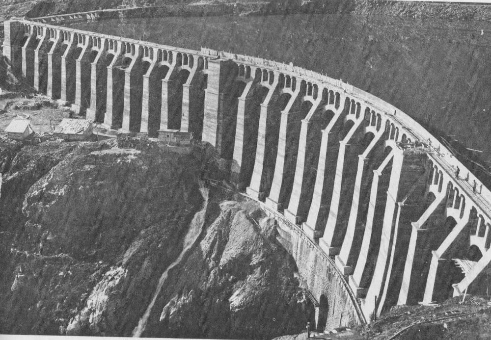 A large, arched concrete dam spans a river in a mountainous area, holding back a large reservoir. Buildings and roads are visible near the base of the dam.