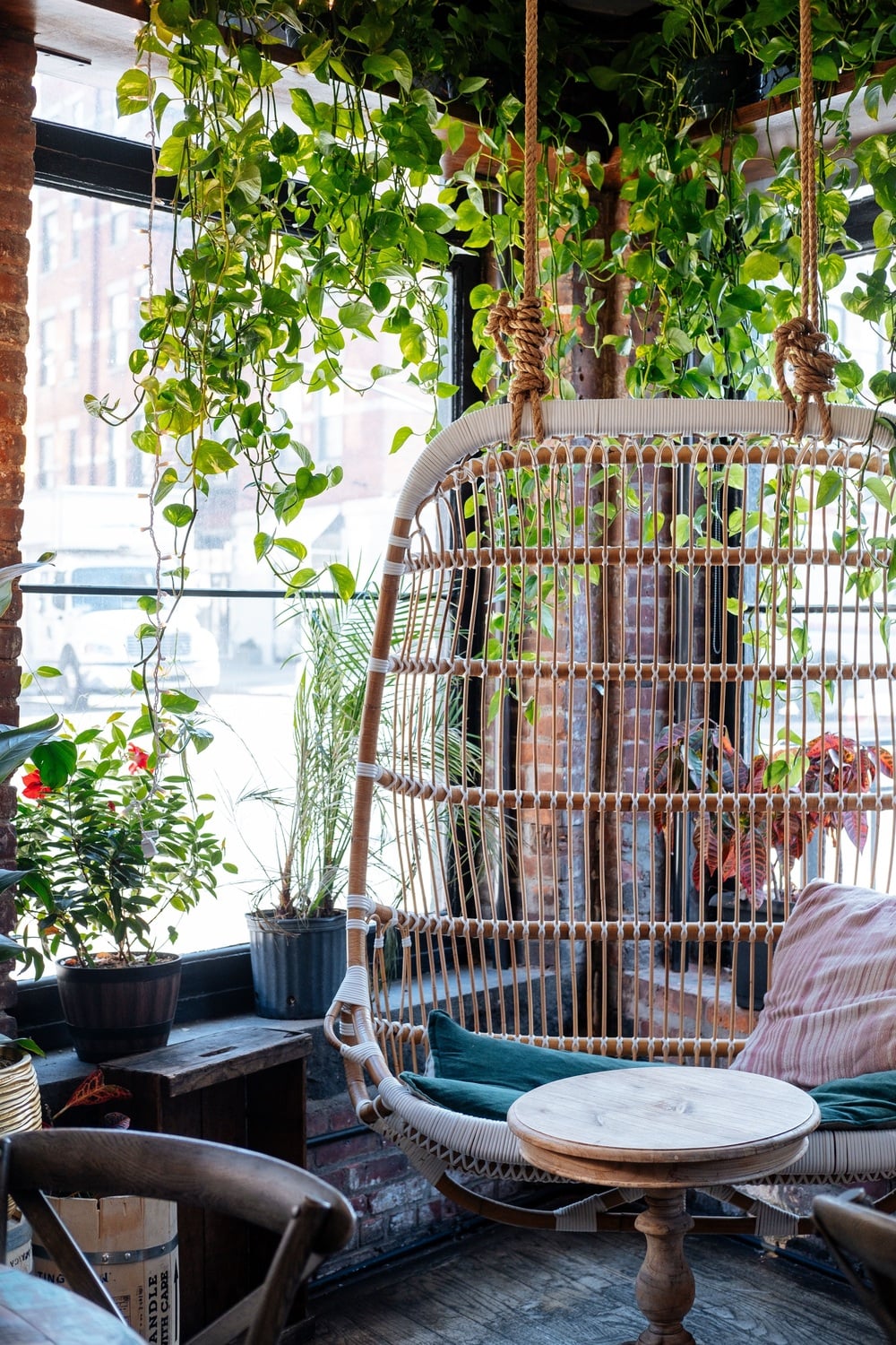 A cozy corner with a hanging wicker chair, cushions, a small wooden table, and various potted plants by large sunlit windows.