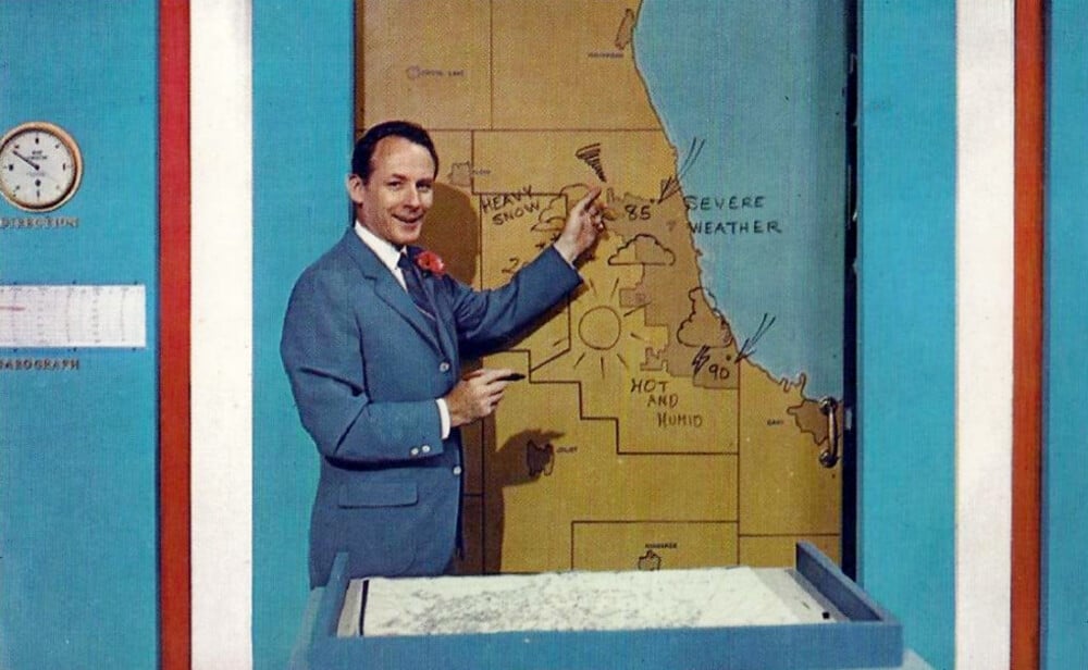 A man in a suit points to a weather map with handwritten notes, showing conditions like “severe weather,” “hot and humid,” and “heavy showers.” There is a clock and calendar on the wall behind him.