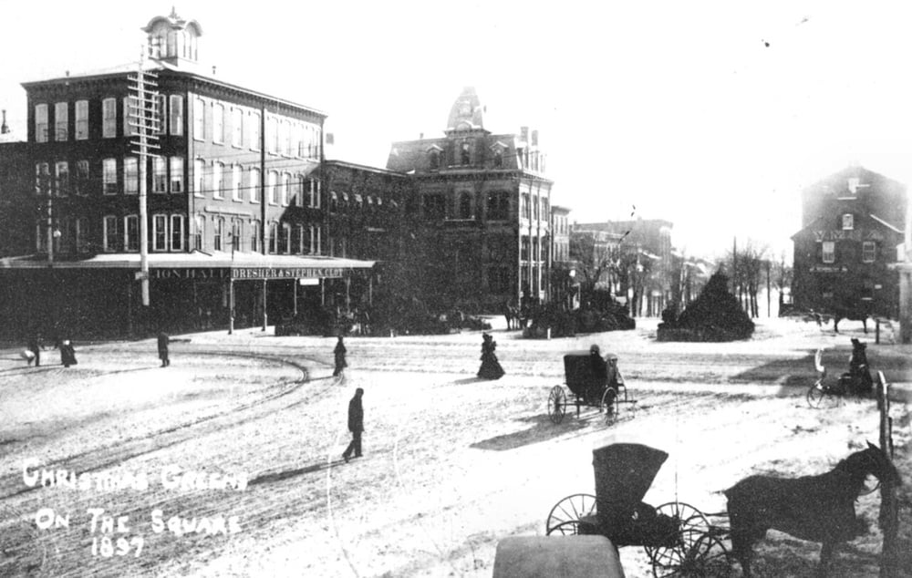 Black and white photo of a snowy town square in 1857, with horse-drawn carriages, people walking, and large brick buildings in the background. Handwritten text reads: “Christmas Greeting on the Square 1857.”