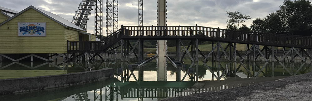 A wooden bridge crosses over a shallow pool near a yellow building labeled “Arcade,” with a tall amusement park ride and cloudy sky in the background.