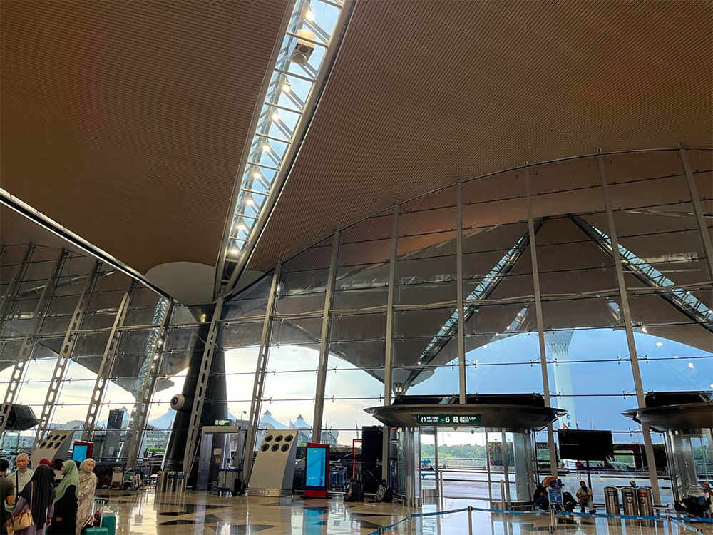 Spacious airport terminal with high arched ceilings, glass walls, and bright natural light. People stand near the entrance; signs and luggage carts are visible in the scene.