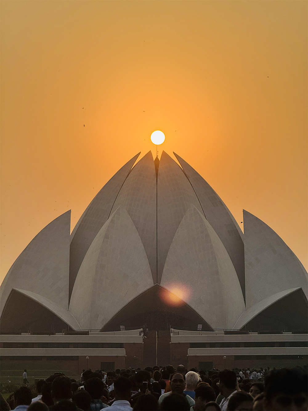 The Lotus Temple at sunset, with the sun appearing to rest perfectly atop the temple’s central spire, surrounded by an orange sky and crowds of visitors in the foreground.