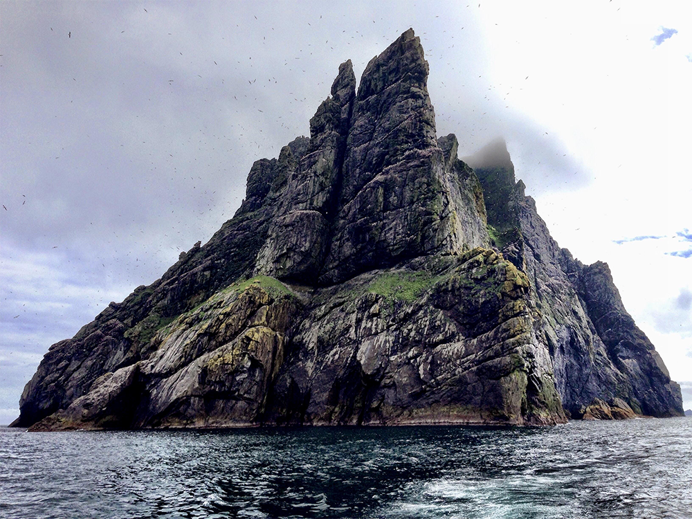 A rocky, steep island rises sharply from the ocean, with patches of green vegetation. The sky is cloudy, and birds are flying around the rugged cliffs. Waves gently lap at the island’s base.