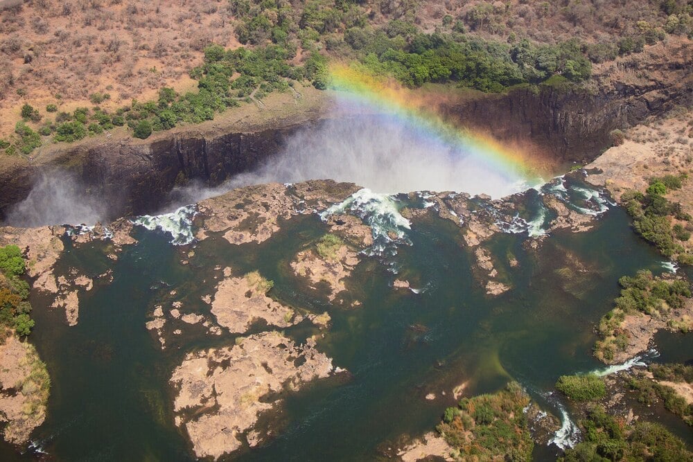 Aerial view of a waterfall with mist rising, a rainbow arching over the falls, and rocky green terrain surrounding the water.