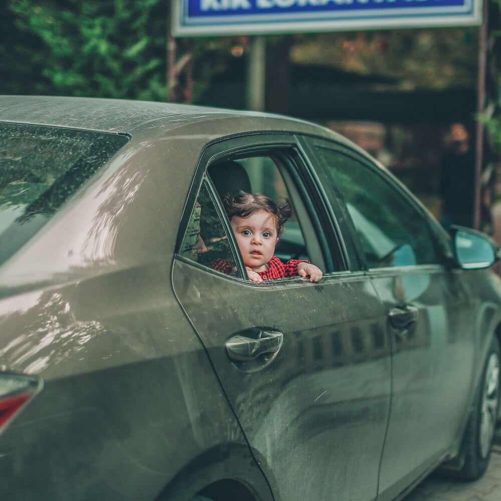 A young child in a red plaid shirt looks out from the back seat window of a parked car on a cloudy day, with greenery and a blurry sign in the background.