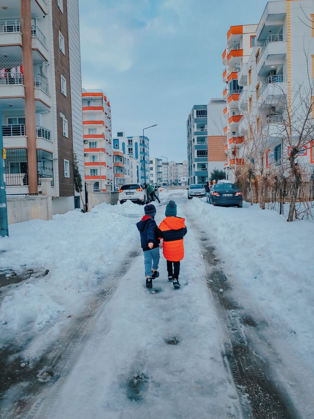 Two children in winter jackets walk down a snowy street lined with apartment buildings and parked cars. Snow covers the ground and sidewalks, creating a winter urban scene.