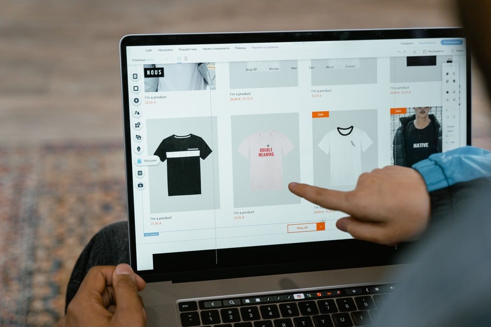 A person points at a laptop screen displaying an online store page with various t-shirts, including a pink one that says “DOUBLE MEANING.”