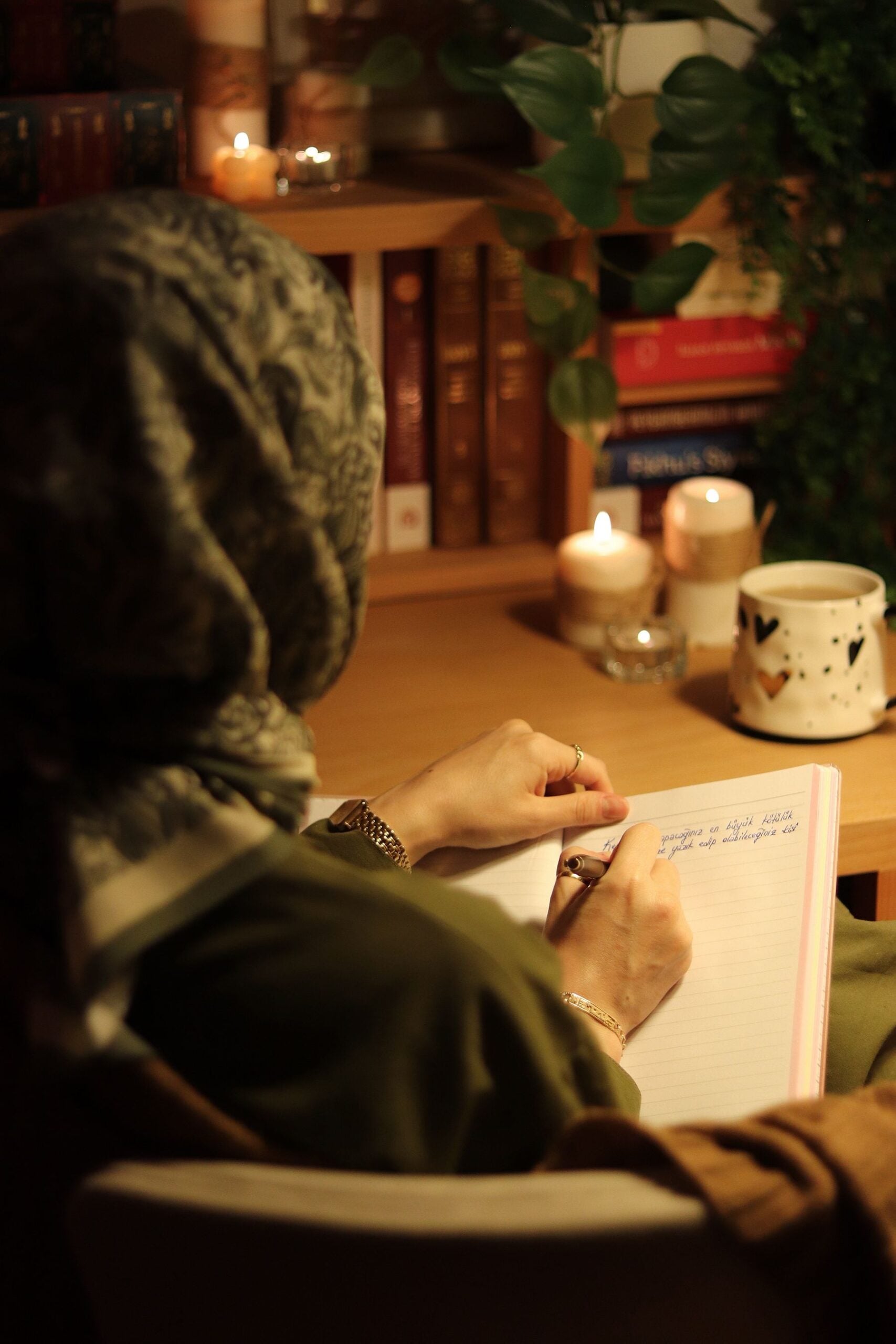 A person wearing a patterned headscarf writes in a notebook at a cozy desk lit by candles, with books and a mug nearby.