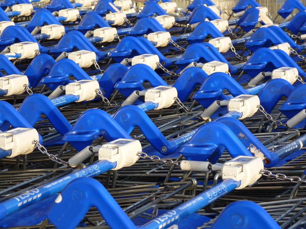 Rows of blue shopping carts with metal frames are neatly lined up outdoors, each cart secured with a locking mechanism and chain. Sunlight highlights the bright blue handles.