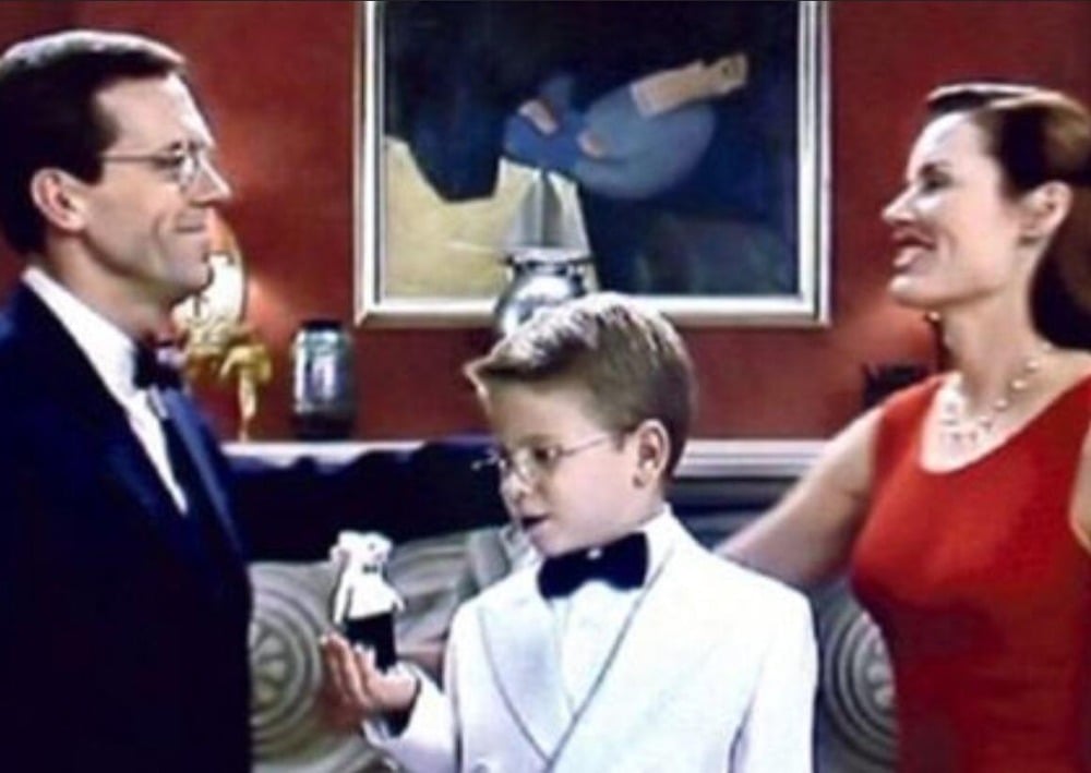 A man, a woman, and a young boy in formal attire stand together indoors, smiling. The boy holds a small white mouse while a colorful painting hangs on the wall behind them.