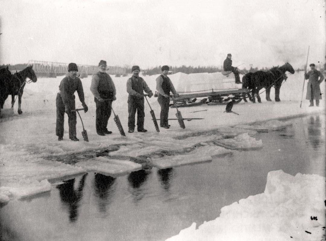Five people stand on snow by an icy river, holding tools. Two horses and a sled are in the background, with another person nearby. The scene appears cold and wintry, with piles of snow and blocks of ice on the ground.