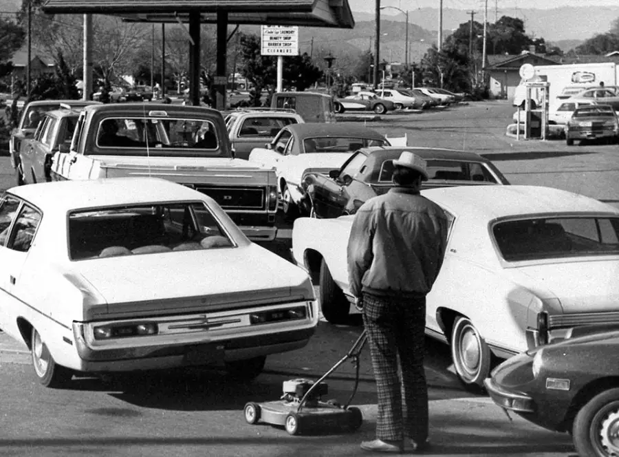 A man with a lawnmower stands in line with cars at a busy gas station, waiting his turn. The scene appears to be from the past, with vintage cars and black-and-white photography.