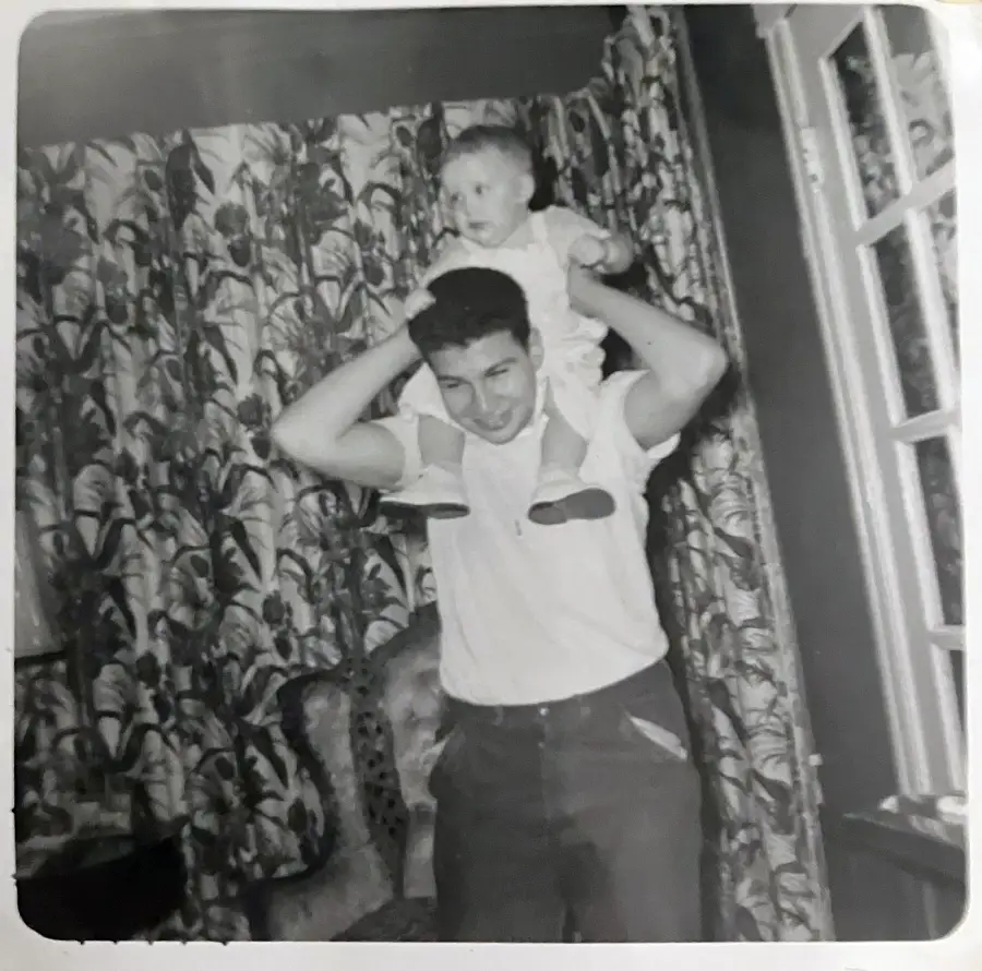 A man in a white T-shirt holds a smiling baby on his shoulders in a room with patterned curtains and a window. Black-and-white photo.