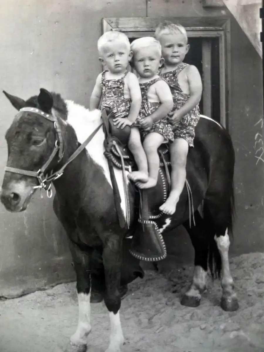 Three young children in matching outfits sit together on a saddled horse outside, posing for a vintage black-and-white photo.