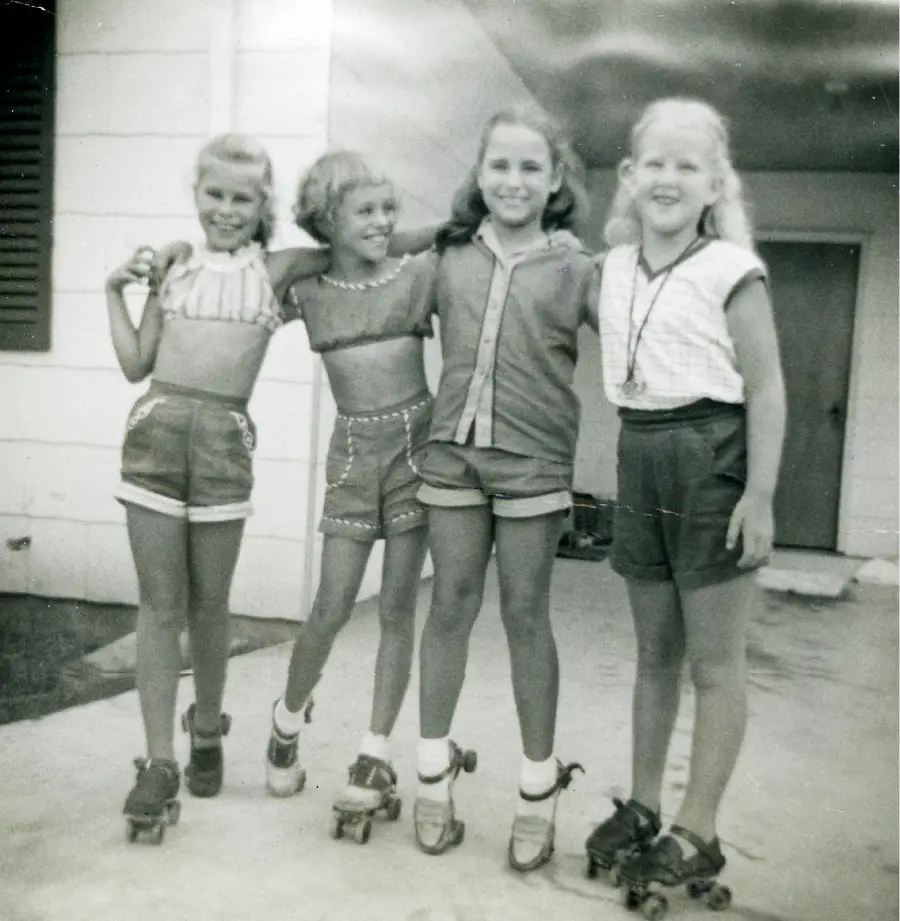 Four young girls stand arm-in-arm outside a building, smiling and wearing roller skates. They are dressed in shorts and summer tops, appearing happy and playful in a black-and-white photo.