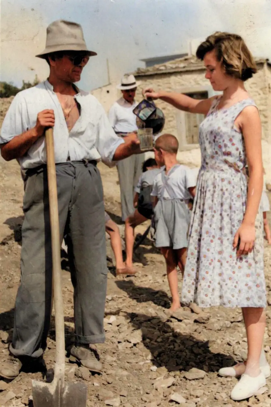 A man with a shovel is handed a drink by a woman in a light dress. Children play in the background near a building on a rocky, sunlit area.