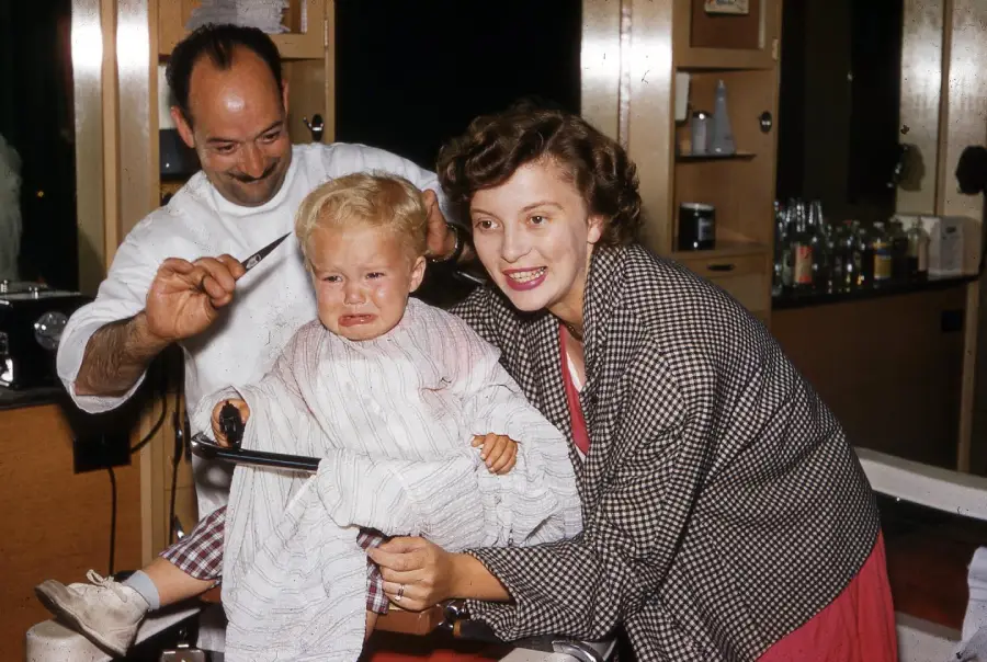 A barber cuts a crying toddler’s hair while a smiling woman, possibly the mother, tries to comfort the child in a bright, vintage barbershop.