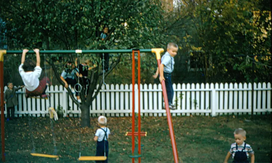 Children play on a swing set and slide in a backyard with a white picket fence and trees in the background. Four kids are visible in the yard, and adults are watching in the background.