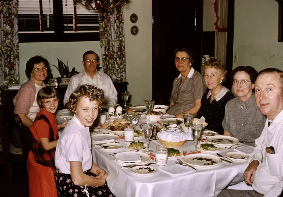 Seven people, including adults and children, sit around a dining table set with dishes and glasses, smiling at the camera in a cozy, warmly lit room with floral curtains.
