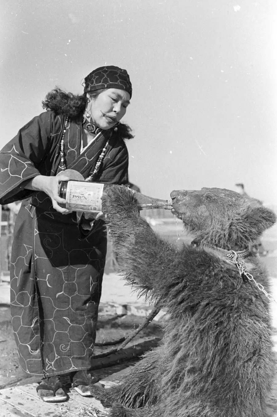 A woman in traditional clothing pours liquid from a bottle into the mouth of a chained bear. The scene appears outdoors, and both are positioned close together, with the woman looking at the bear. Black and white photo.