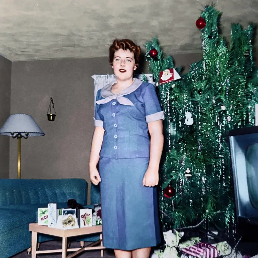 A woman in a blue dress stands in a living room beside a tinsel Christmas tree with ornaments and gifts. A TV, cards on a table, and a sofa are also visible in the background.