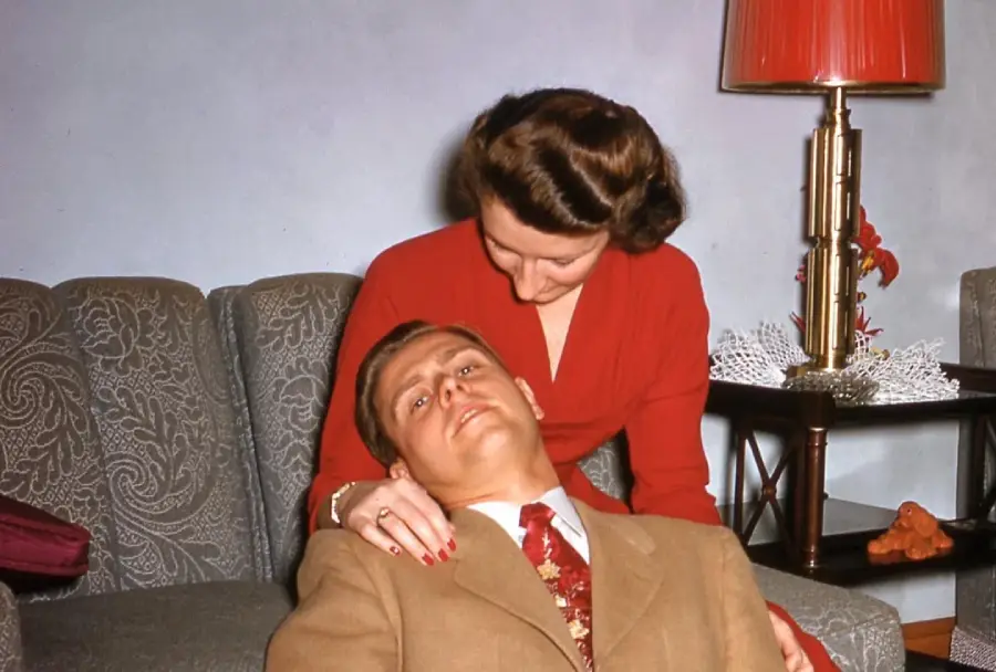 A woman in a red dress holds a man in a brown suit, who is lying with his head in her lap on a patterned couch. A red lamp and a doily are on a side table behind them.