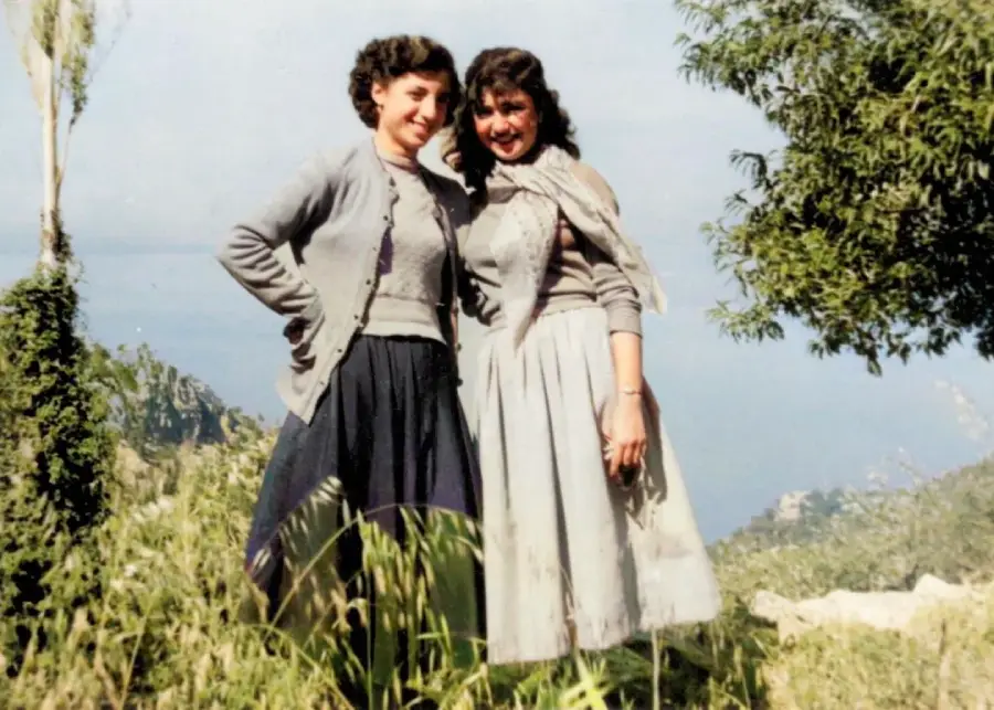 Two women in mid-century clothing stand close together, smiling outdoors on a grassy hillside with green trees and a hazy sky in the background.