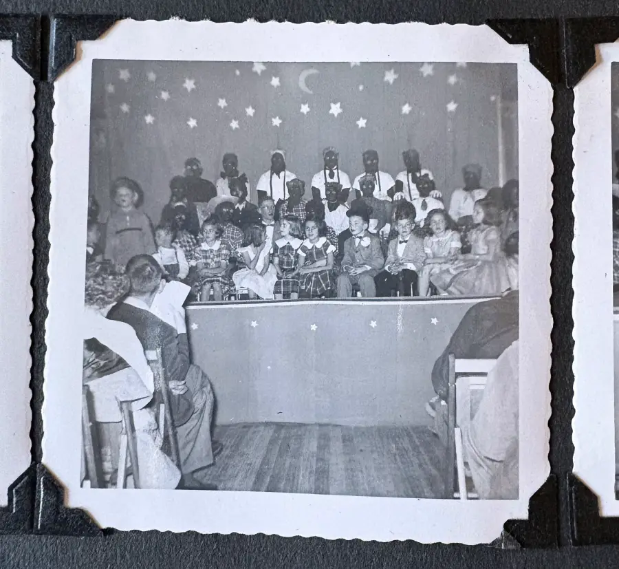 Black and white photo of children and adults on stage in costume, with an audience in front. The backdrop has stars and a moon, suggesting a night or celestial theme.