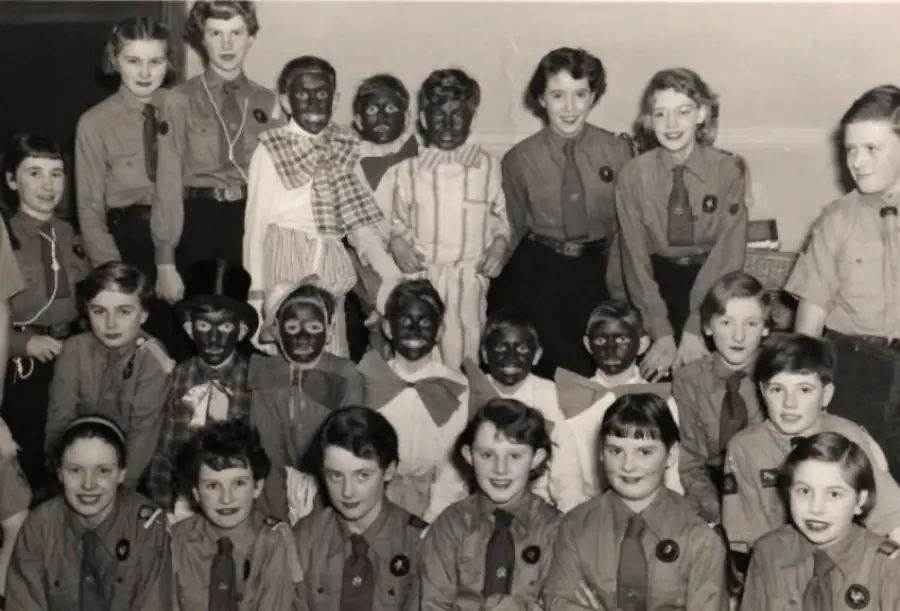 A black-and-white photo of a group of girls, some in scout uniforms and several in blackface makeup, posing and smiling for the camera.