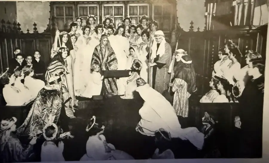 A black-and-white photo of a large group in formal medieval-style costumes, gathered in a wood-paneled room; some are seated, others stand around a central figure, suggesting a staged performance or ceremony.