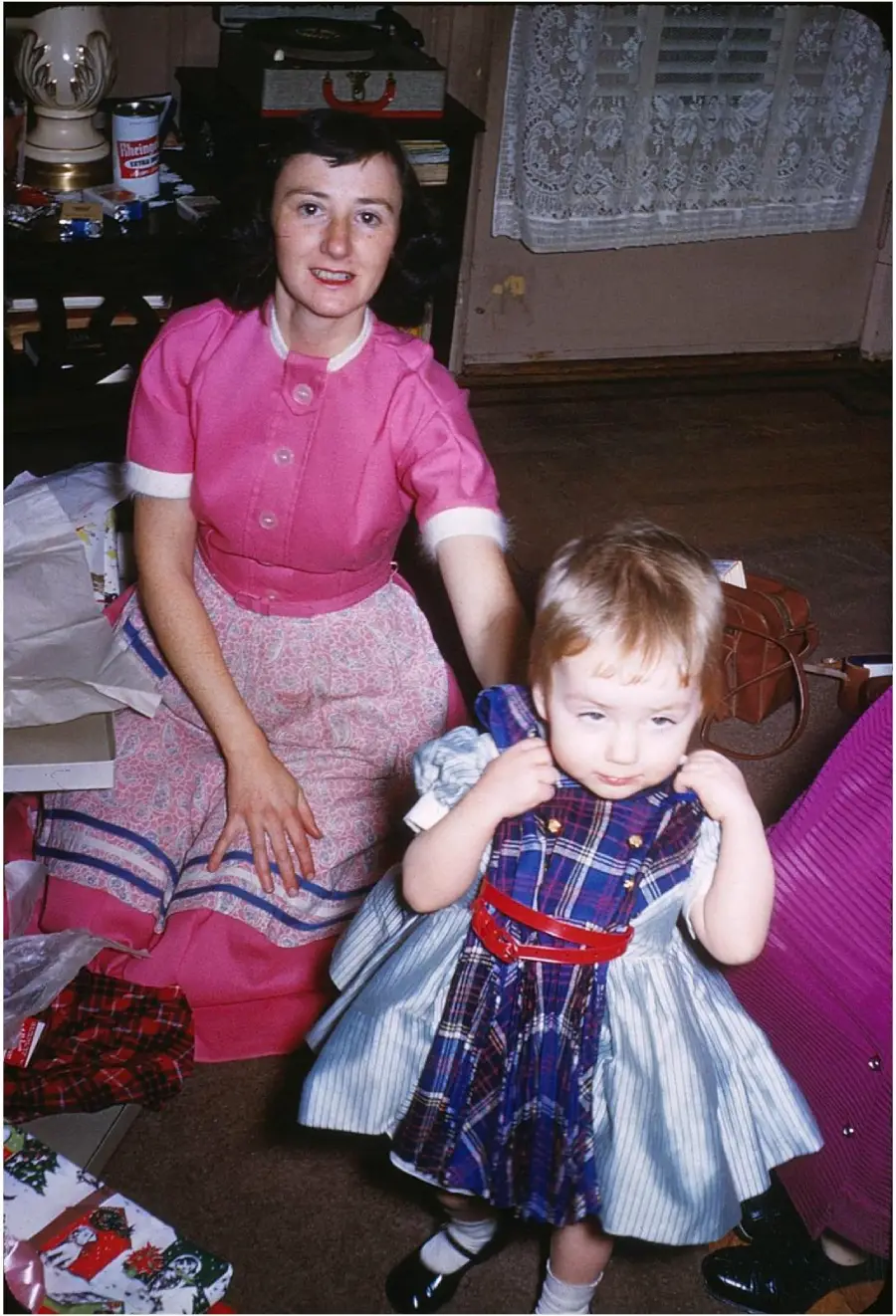 A woman in a pink dress sits on the floor next to a young girl in a plaid dress. They are indoors, possibly celebrating a holiday, with gifts and wrapping paper around them.