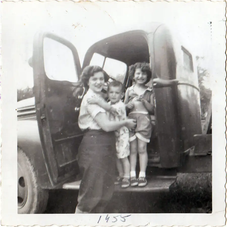 A woman stands by an old truck, holding a young boy, with a smiling girl standing in the truck’s doorway. The photo is black and white and dated 1955.