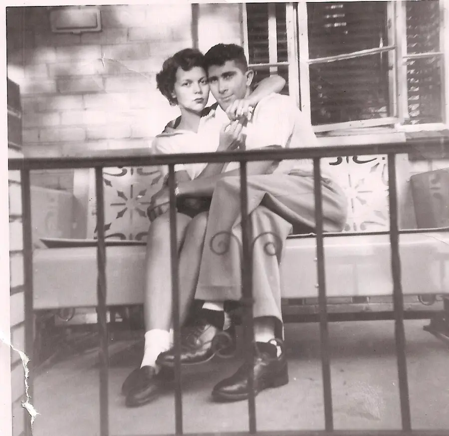 A black-and-white photo of a young couple sitting closely on a porch swing, with the man’s arm around the woman and a metal railing visible in the foreground.