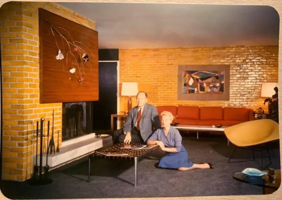 A man in a suit and a woman in a blue dress kneel by a coffee table in a mid-century modern living room with brick walls, abstract art, and stylish furniture.