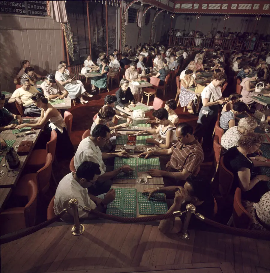 A large group of people sit at tables in a hall, focused on playing bingo with cards and markers. The room is crowded, with participants of various ages under warm lighting. Velvet ropes separate the audience from the main area.
