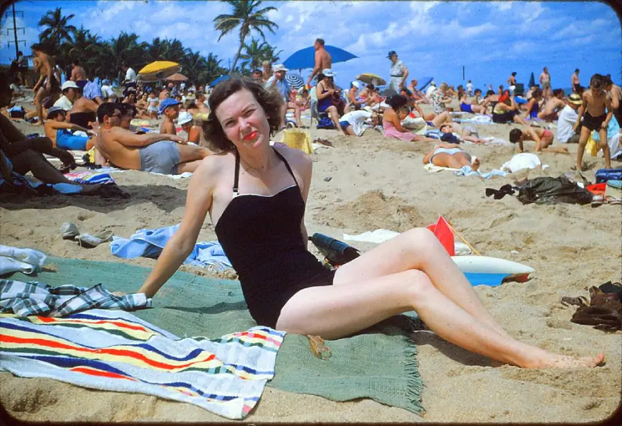 A woman in a black swimsuit sits on a towel at a crowded beach, smiling at the camera. People around her sunbathe and relax under umbrellas. Palm trees and blue sky are visible in the background.