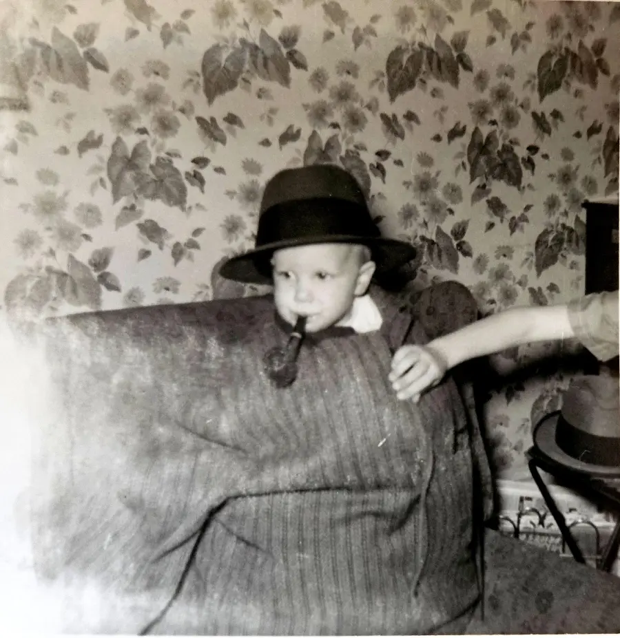 A young child wears an oversized coat and hat, holding a pipe in their mouth, sitting in front of floral wallpaper. An arm reaches toward the child from the right side of the image.