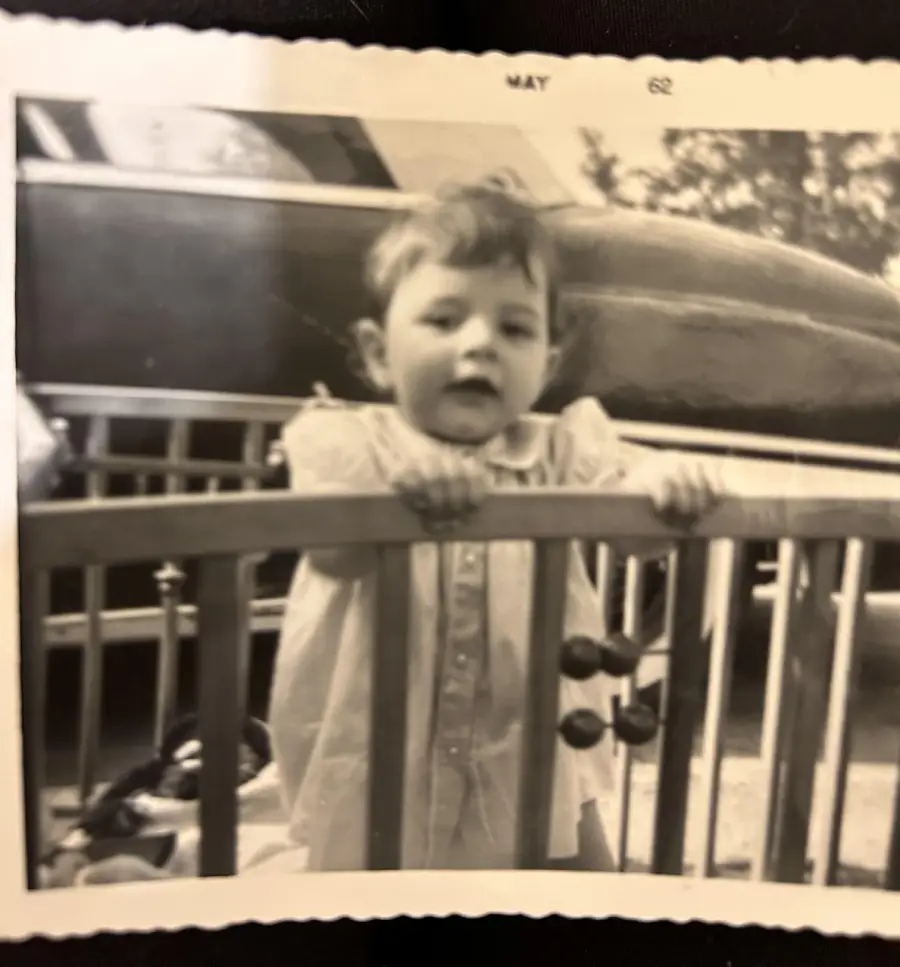 A black-and-white photo from May 1962 shows a baby standing in a crib, holding onto the wooden rail. The baby is wearing a light-colored outfit, and there is a blurred background with a car and trees visible.